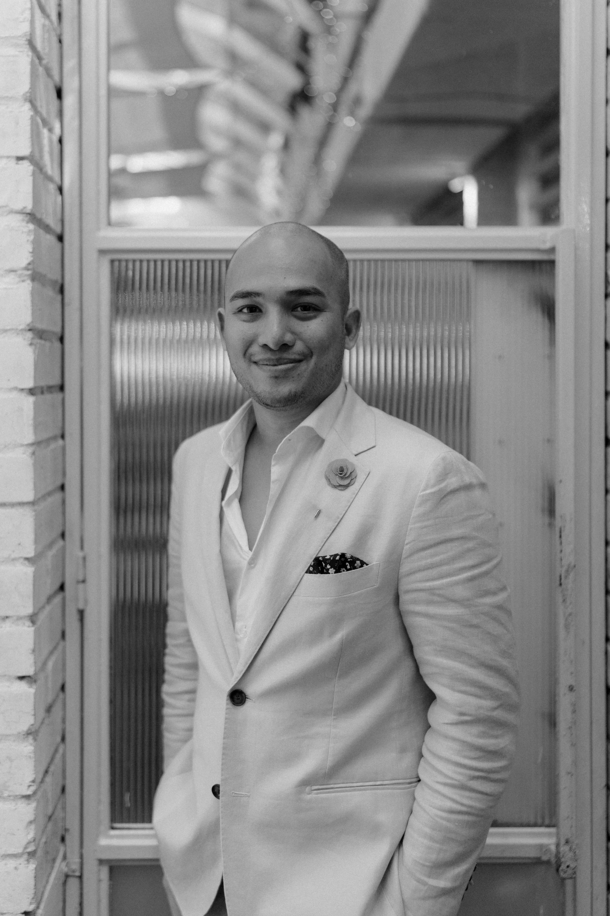 A smiling man in a light suit with a flower lapel pin, standing in front of a textured glass window, with a brick wall on the side.