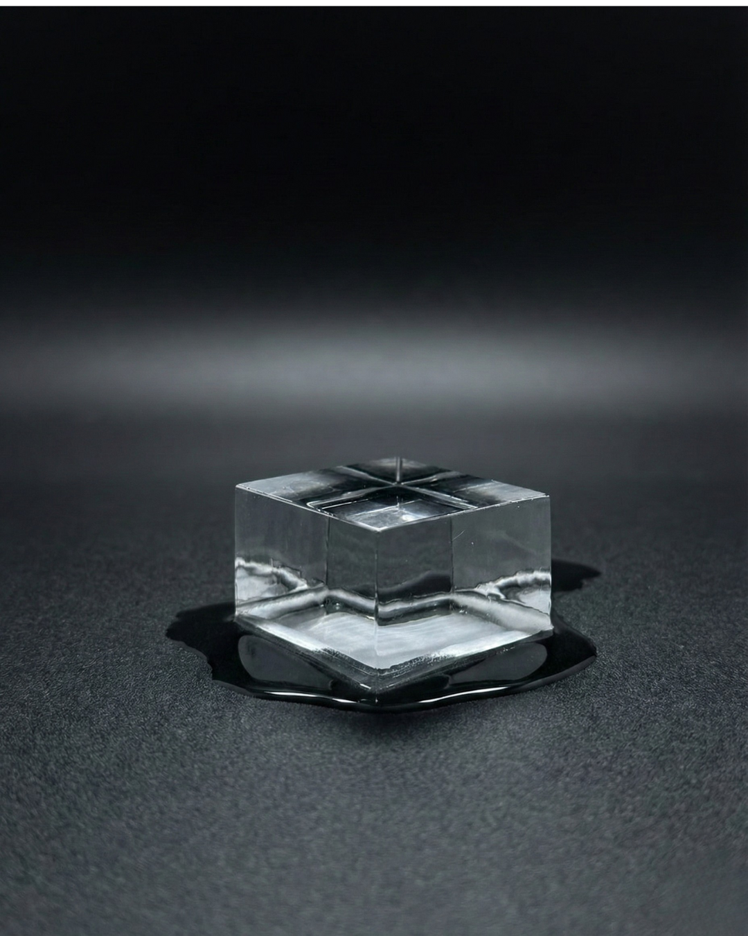 A single clear ice cube melting on a dark surface with a small pool of water around it, against a black background.