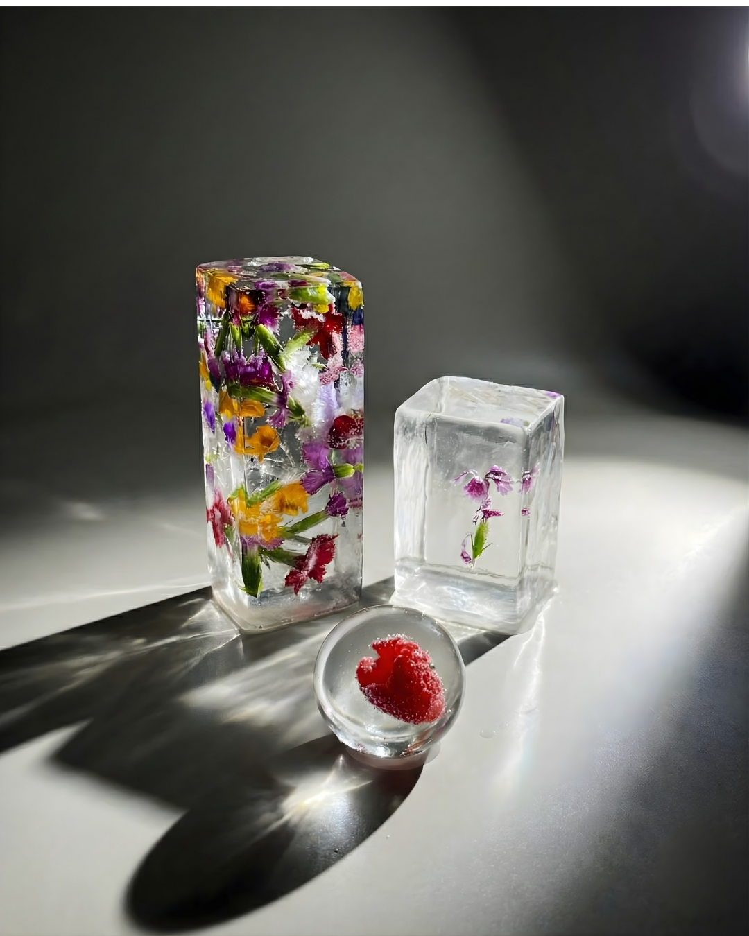 Three ice blocks, one tall block with embedded colorful flower petals, one clear cube with purple flowers inside, and one small round ice with a red fruit frozen inside, placed on a gray surface with shadows.