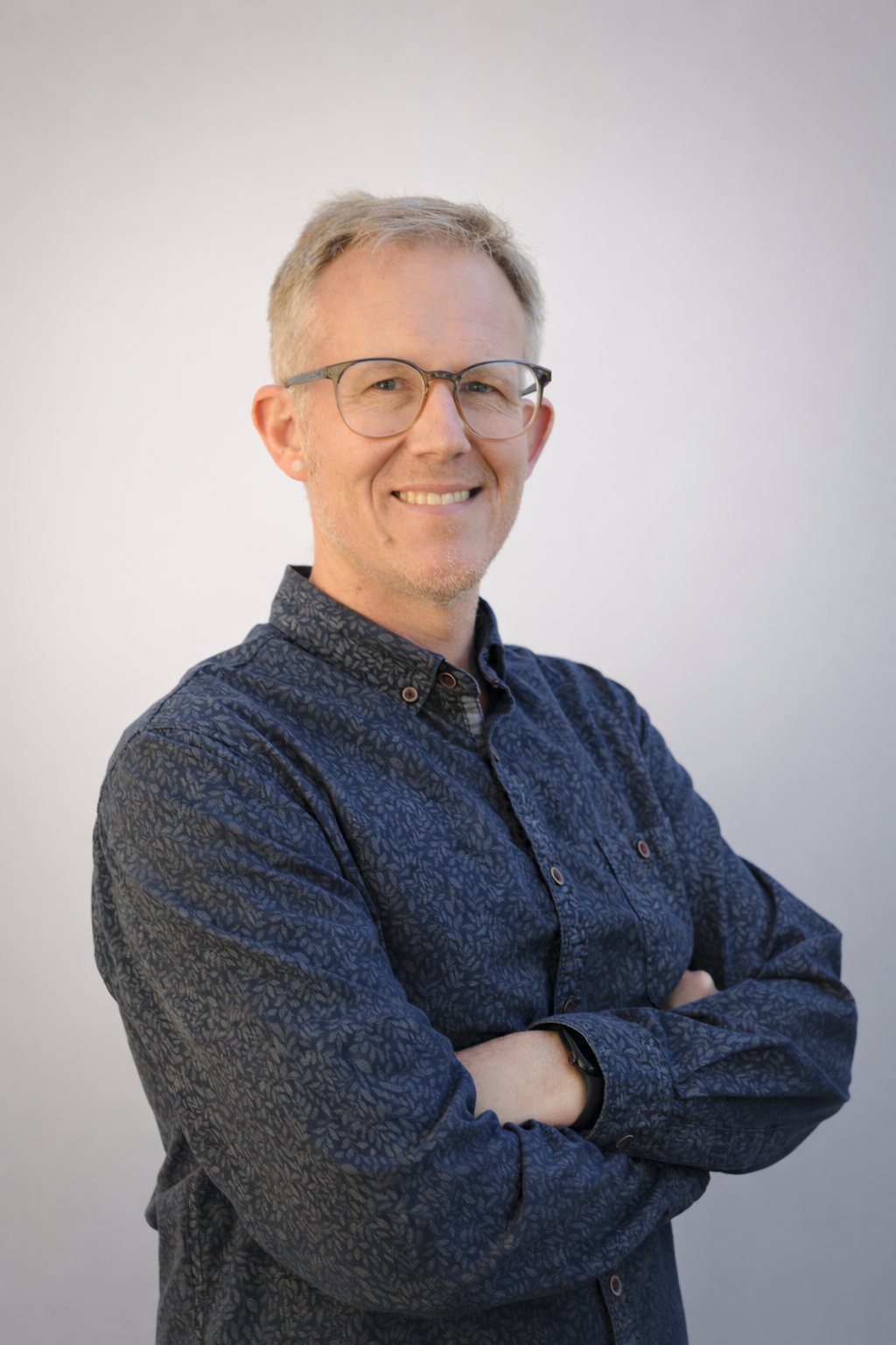 A man with glasses and light brown hair, smiling with arms crossed, wearing a dark patterned shirt against a plain background.