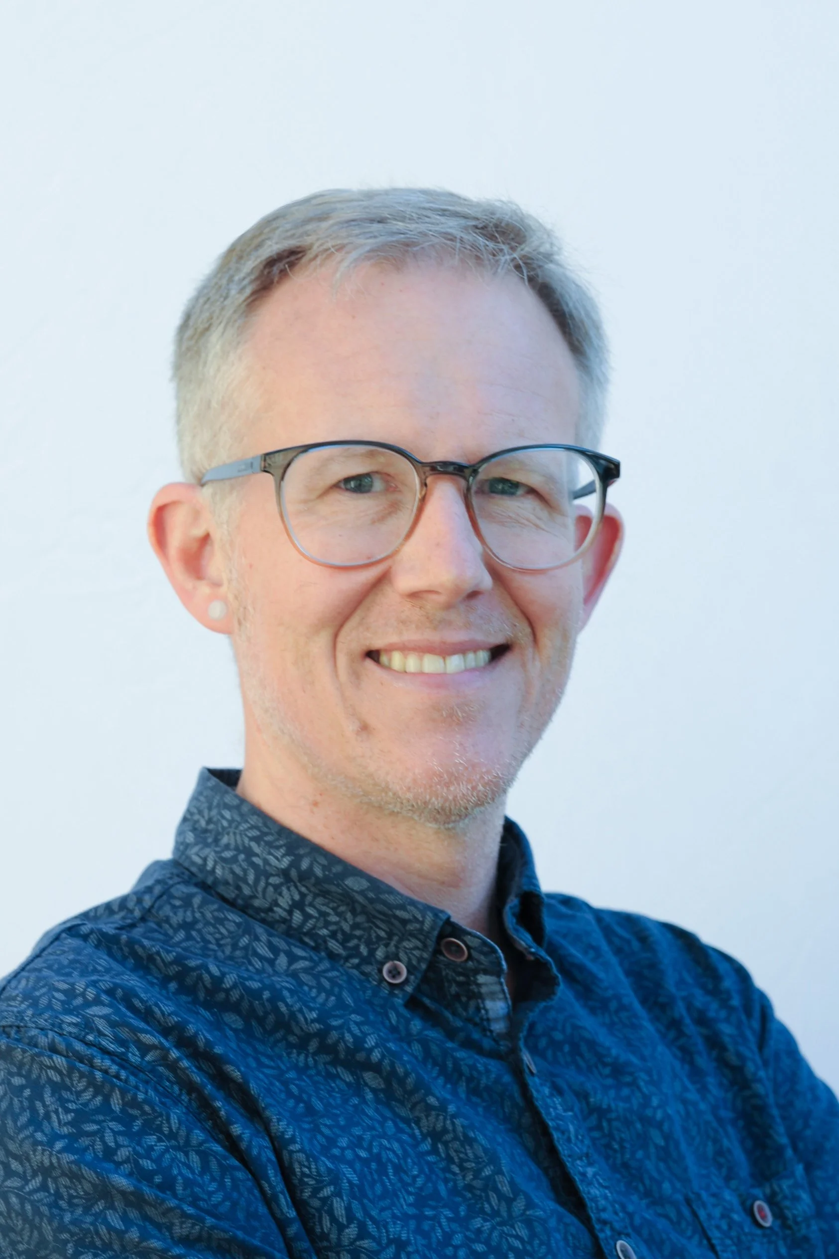 Portrait of a man smiling, wearing glasses and a patterned dark blue shirt, standing against a plain light-colored background.