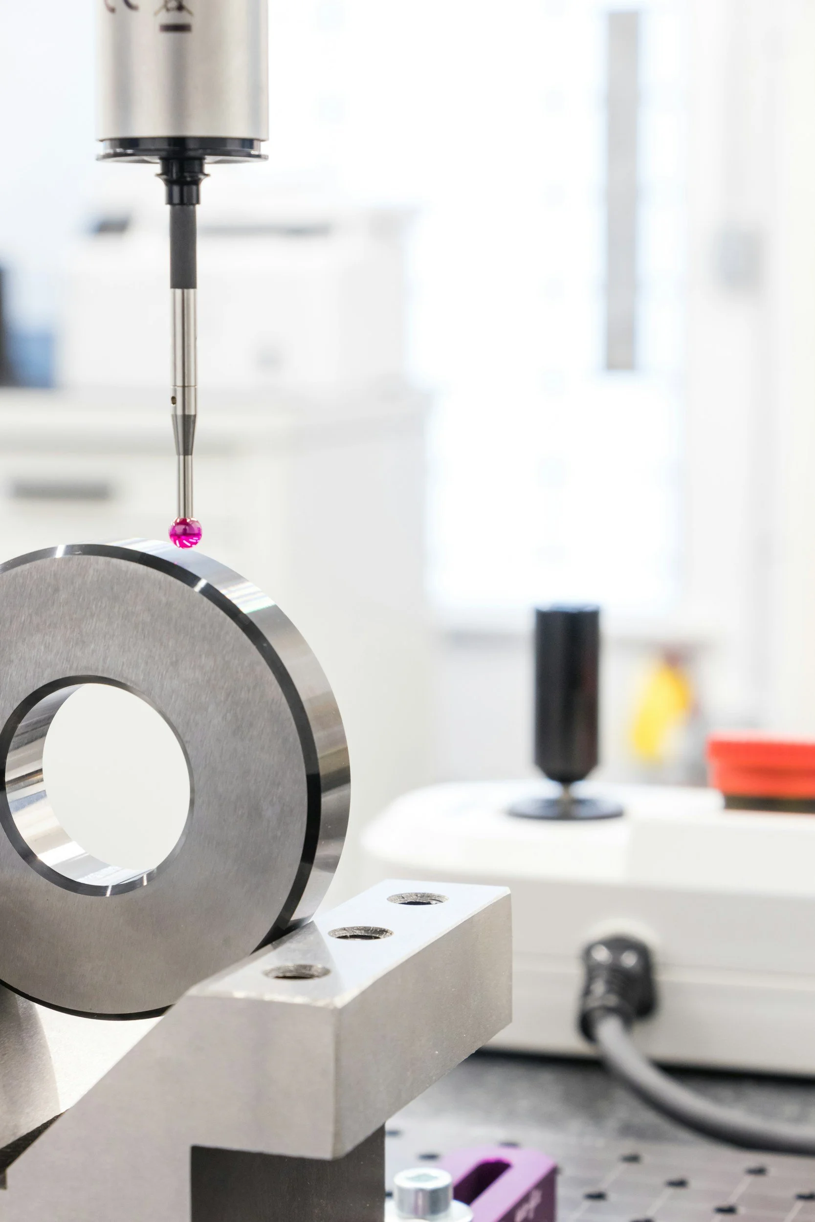 Laboratory setup with a metallic circular component held in place, a pin with a pink bead, and scientific equipment in the background.
