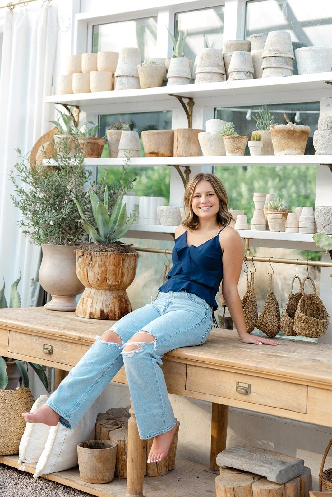 A smiling young woman with shoulder-length brown hair, wearing a navy sleeveless top and ripped jeans, sitting on a wooden table in a bright space filled with potted plants and decorative gardening items.