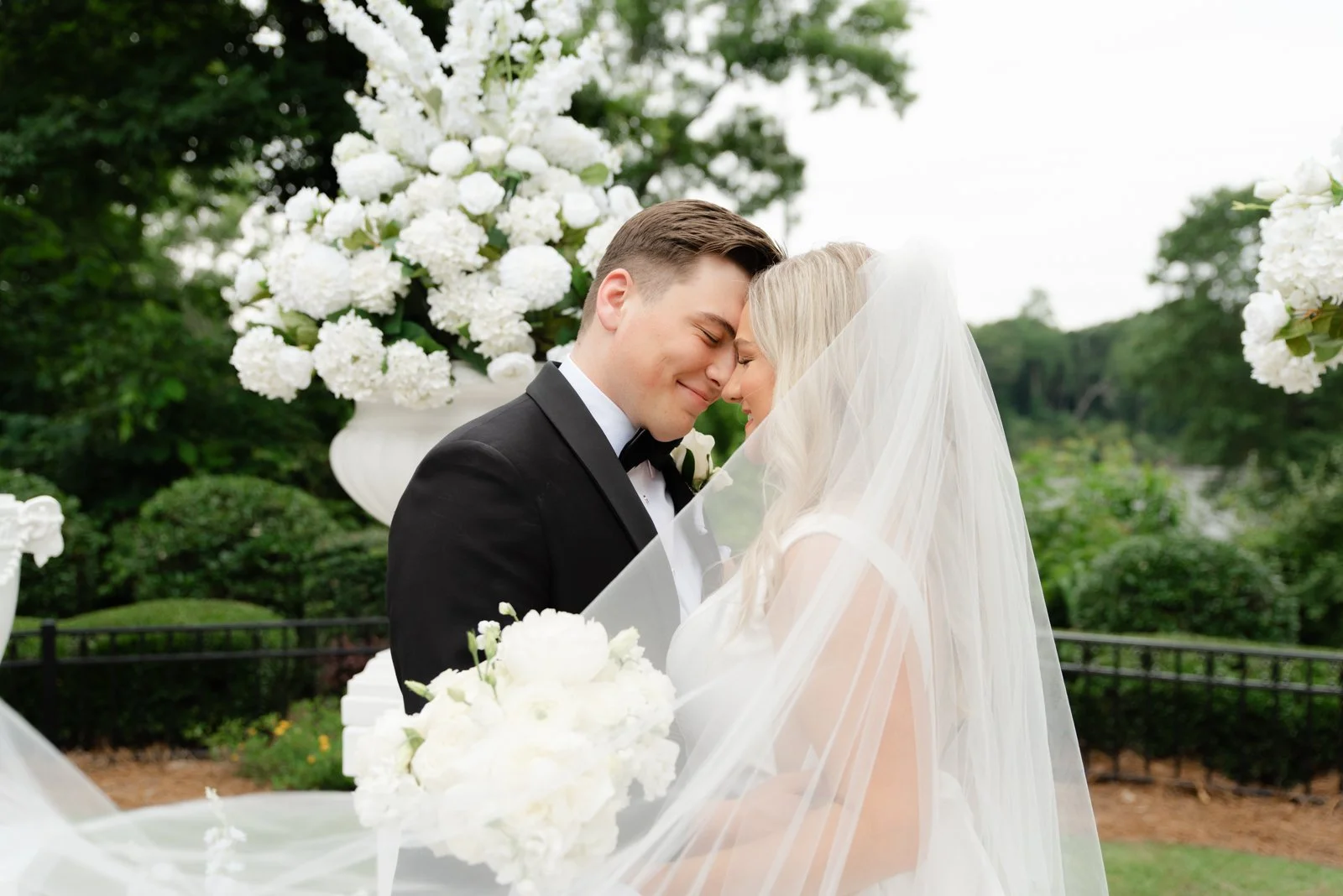 A bride and groom sharing a moment with foreheads touching, dressed in wedding attire, outdoors with white floral arrangements and greenery in the background.