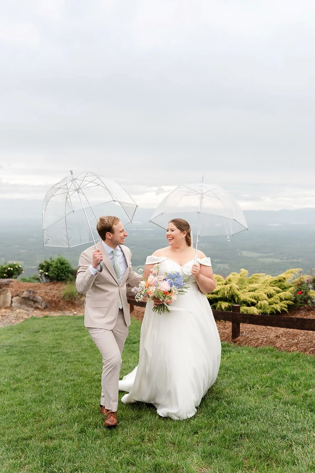 A bride and groom holding clear umbrellas, walking on grass with a scenic view behind them, smiling and looking at each other.