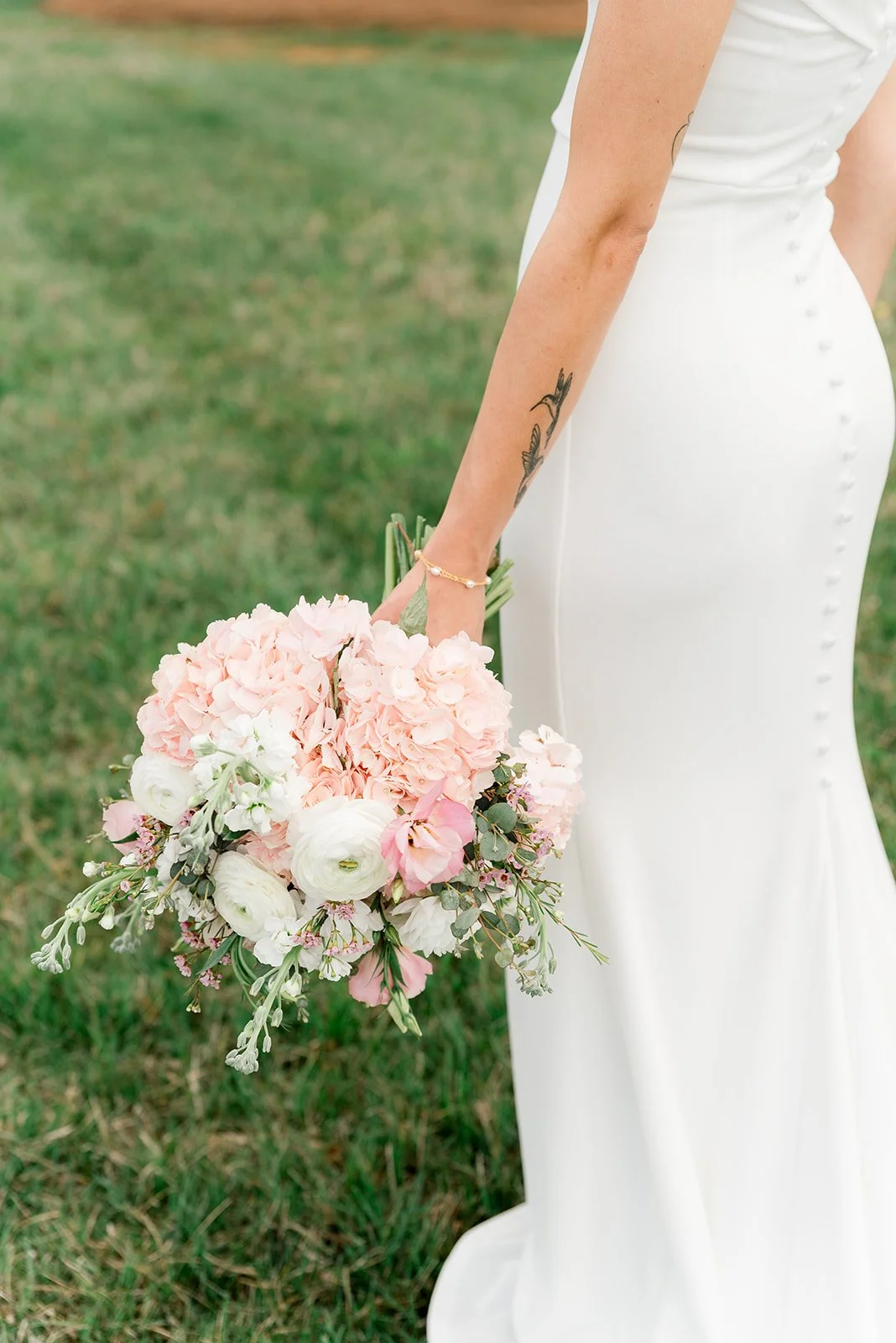Close-up of a bride holding a bouquet of pink and white flowers outdoors, wearing a white wedding dress and a gold bracelet.