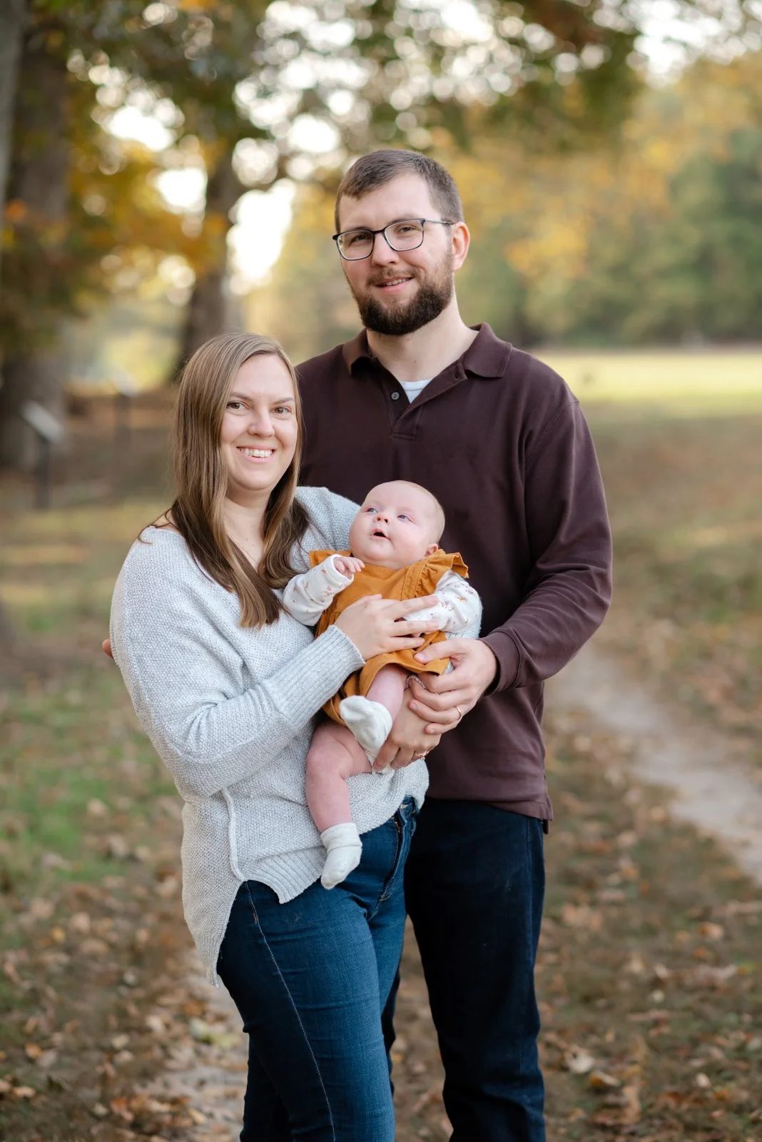 A young family of three stands outdoors on a fall day, with trees and leaves in the background. The woman holds a baby girl, and the man stands next to them, smiling.