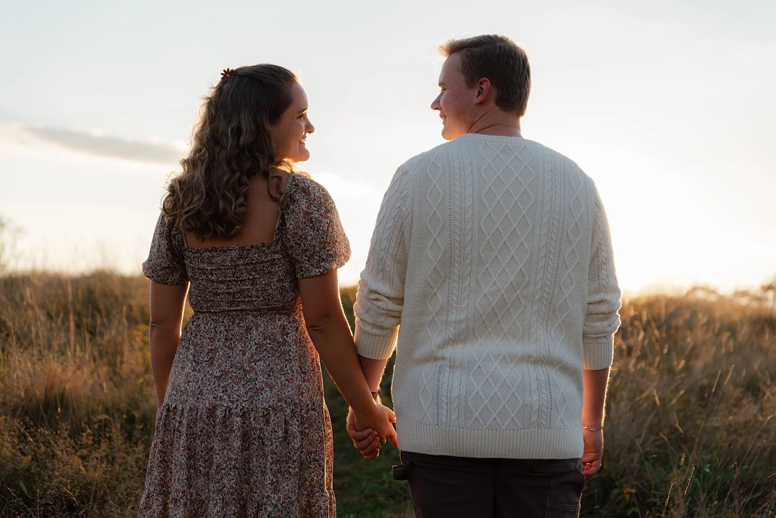 A couple holding hands, standing in a grassy field during sunset, facing each other and smiling.