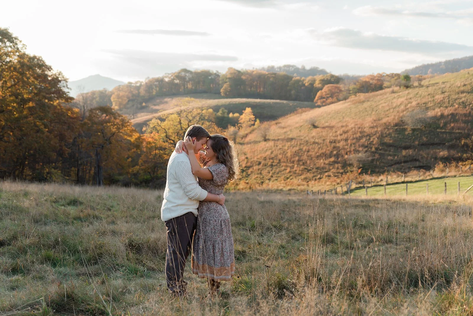 A couple embracing in a field with hills and trees in autumn foliage in the background during sunset.