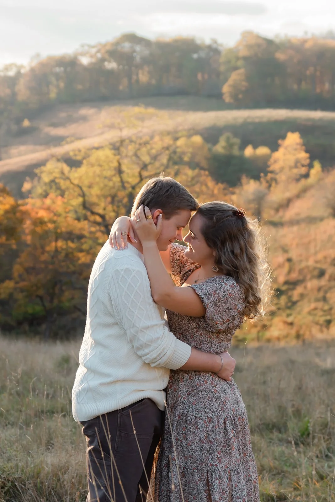 A couple embracing outdoors during sunset with autumn trees and rolling hills in the background.