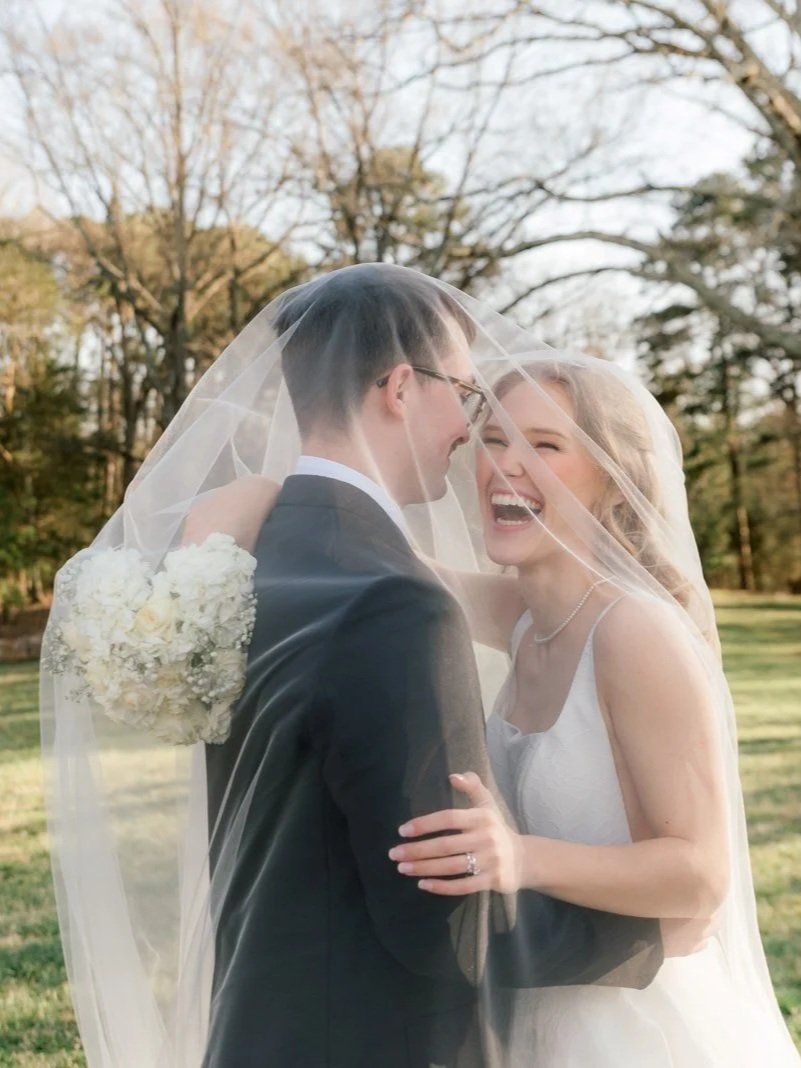 A bride and groom sharing a joyful moment outdoors, with the bride's veil draped over both of them, and a bouquet of white flowers in the background.