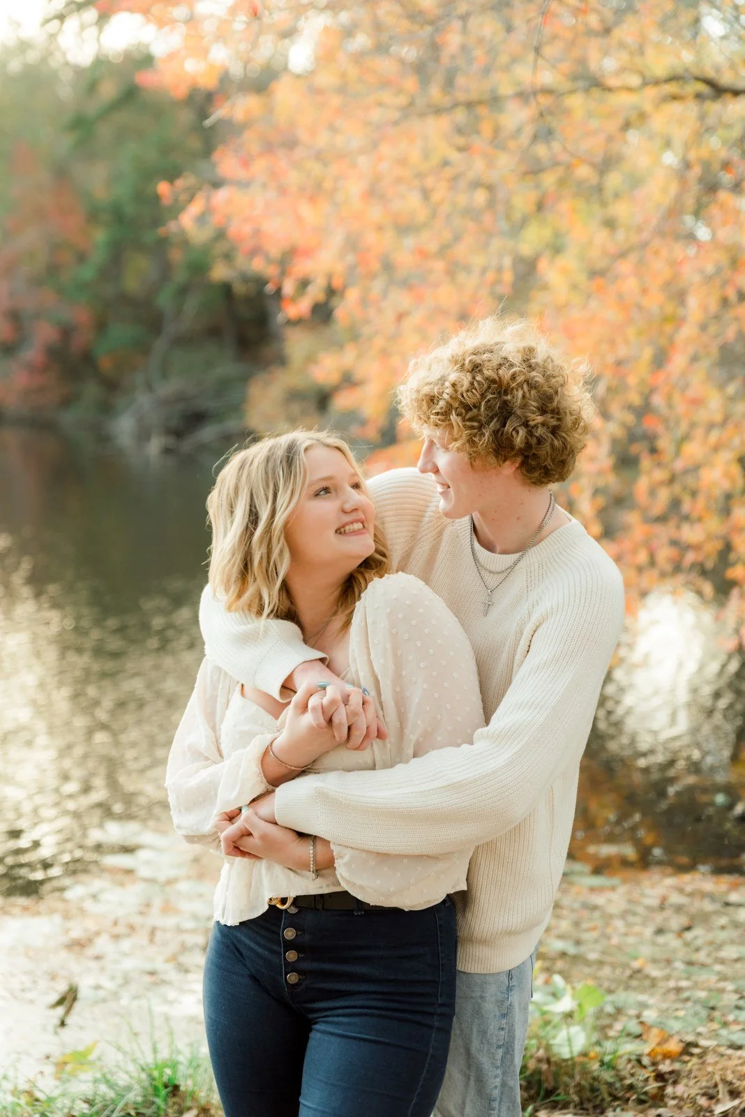 A young couple smiling and embracing outdoors by a river in autumn, with colorful fall leaves in the background.