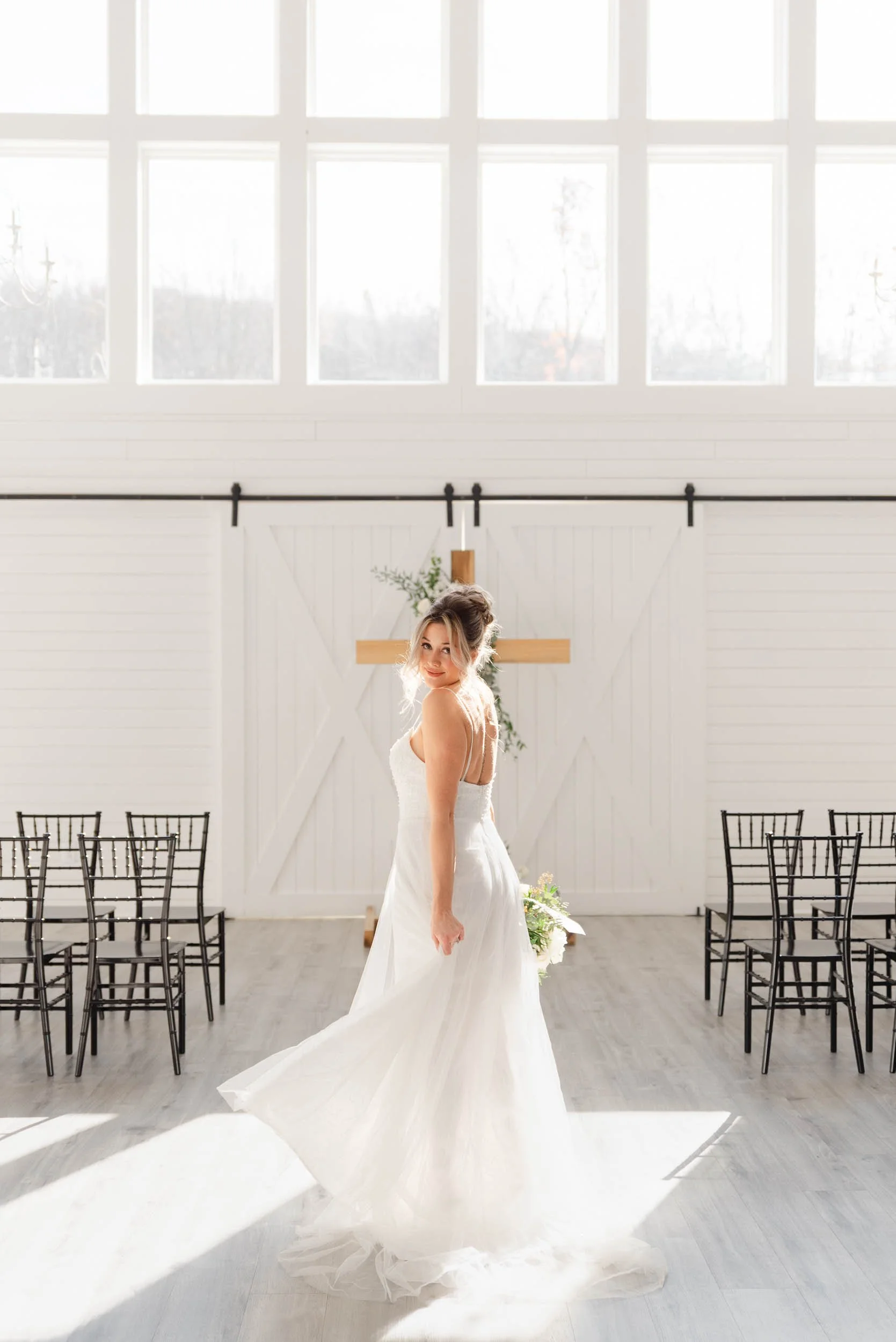 A bride in a white wedding dress standing in a bright, modern barn-style venue with white walls, large windows, and black chairs. She is looking over her shoulder with sunlight streaming in.
