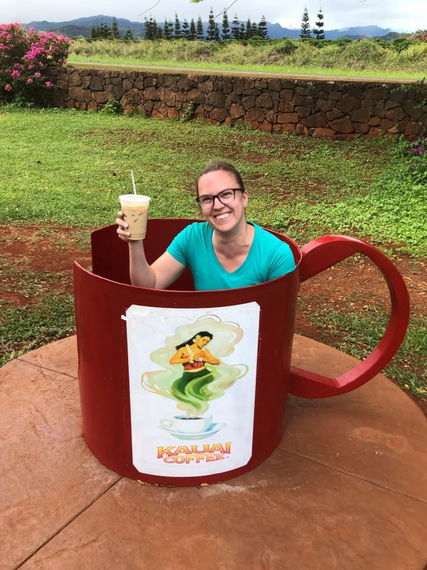 A woman smiling and sitting in a large red coffee mug-shaped structure outdoors, holding a plastic cup of iced coffee. The mug has a sign on it that says 'Kauai Coffee' with an illustration of a hula dancer. The background includes green grass, a stone wall, and some trees.