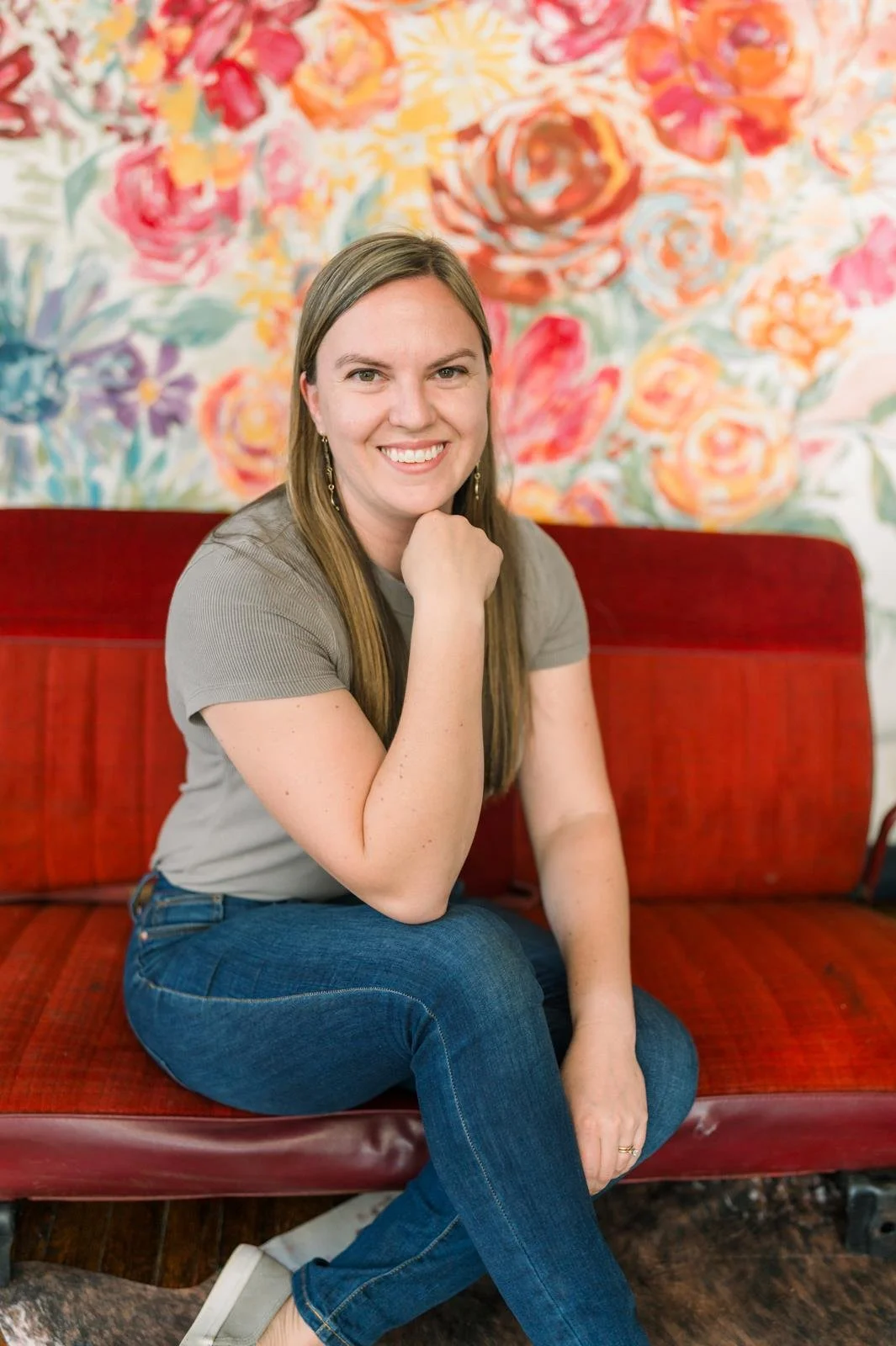 A woman with long brown hair and a gray shirt sitting on a red bench in front of a colorful floral painting.
