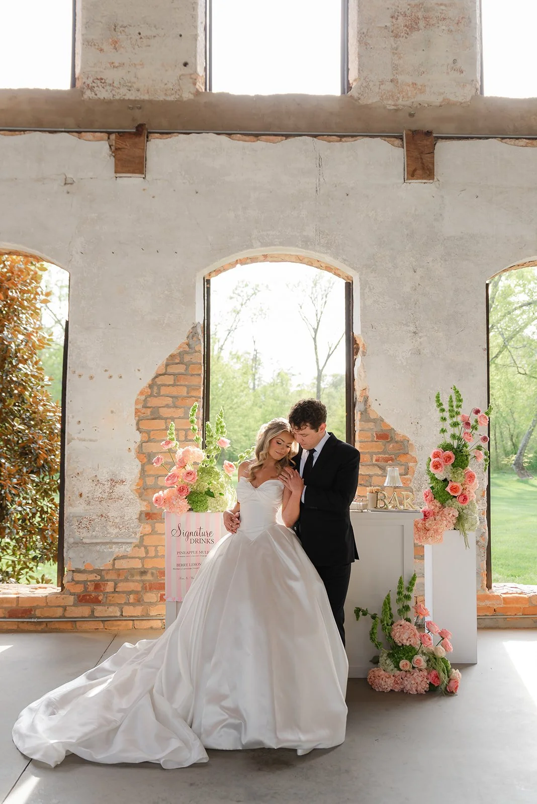 A bride and groom sharing a romantic moment indoors, with a background of large, open brick and concrete windows, and decorated with pink and green floral arrangements.