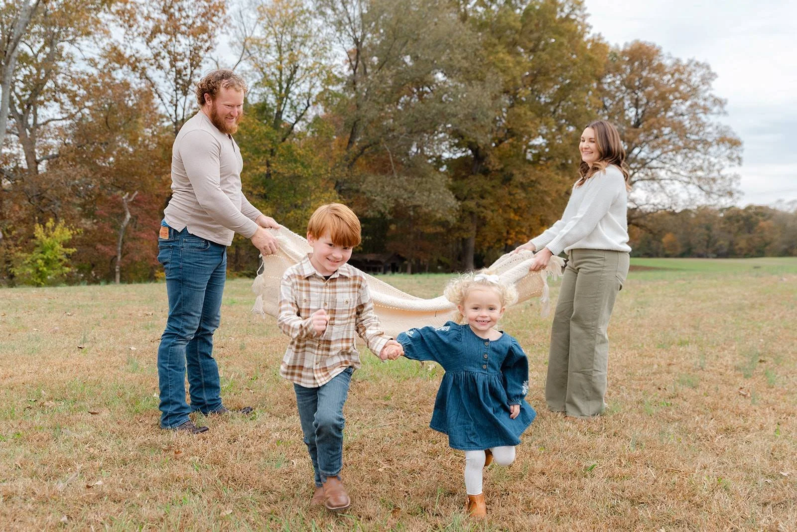 Two adults and two children playing in a park with trees showing autumn colors, holding a blanket and smiling.