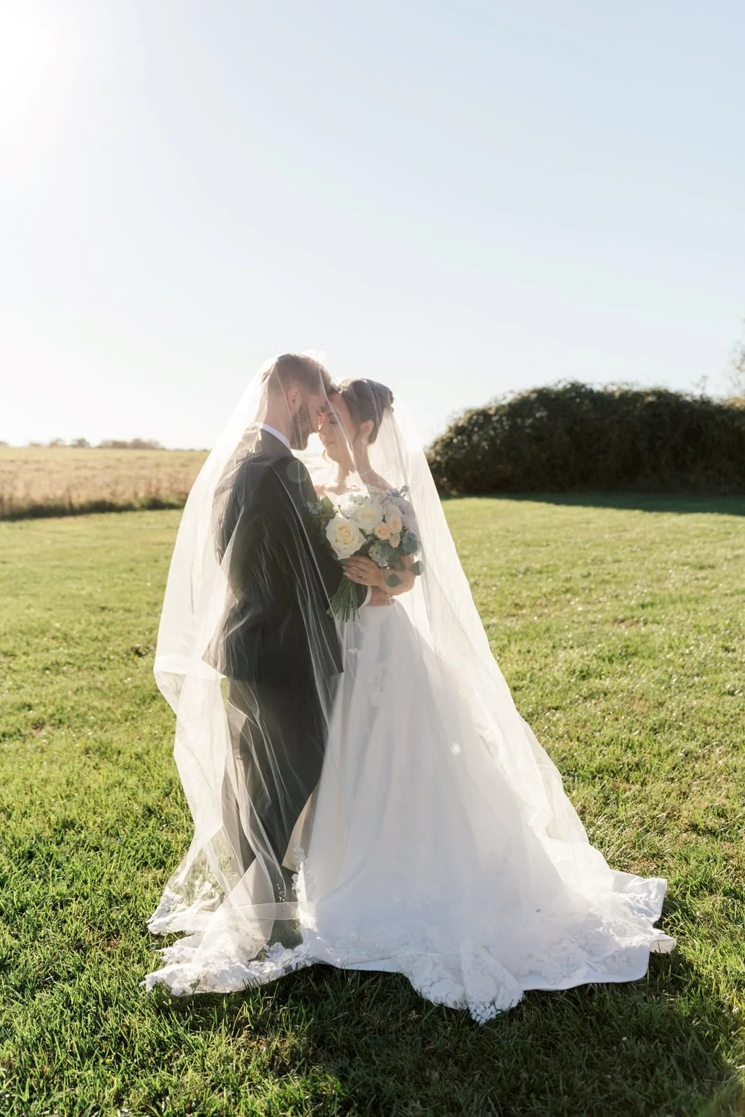 A bride and groom standing under a veil in a grassy field at sunset, holding a bouquet of white roses and facing each other with foreheads touching.