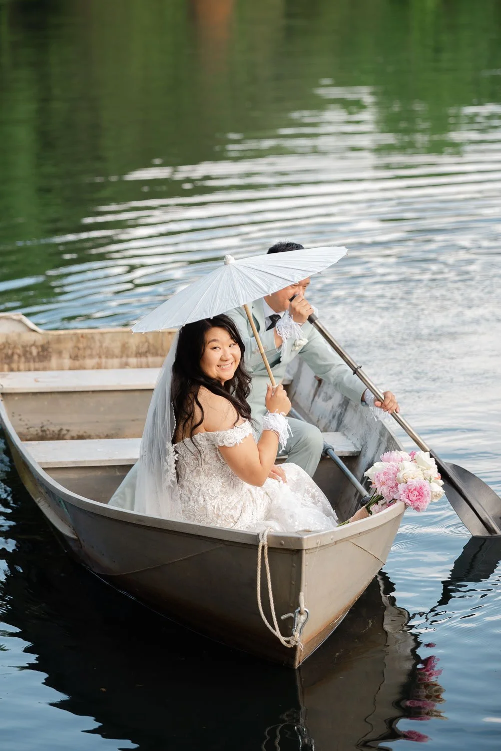 A bride and groom in wedding attire sitting in a small boat on a calm body of water, with the bride holding a white parasol and a bouquet of pink and white flowers.