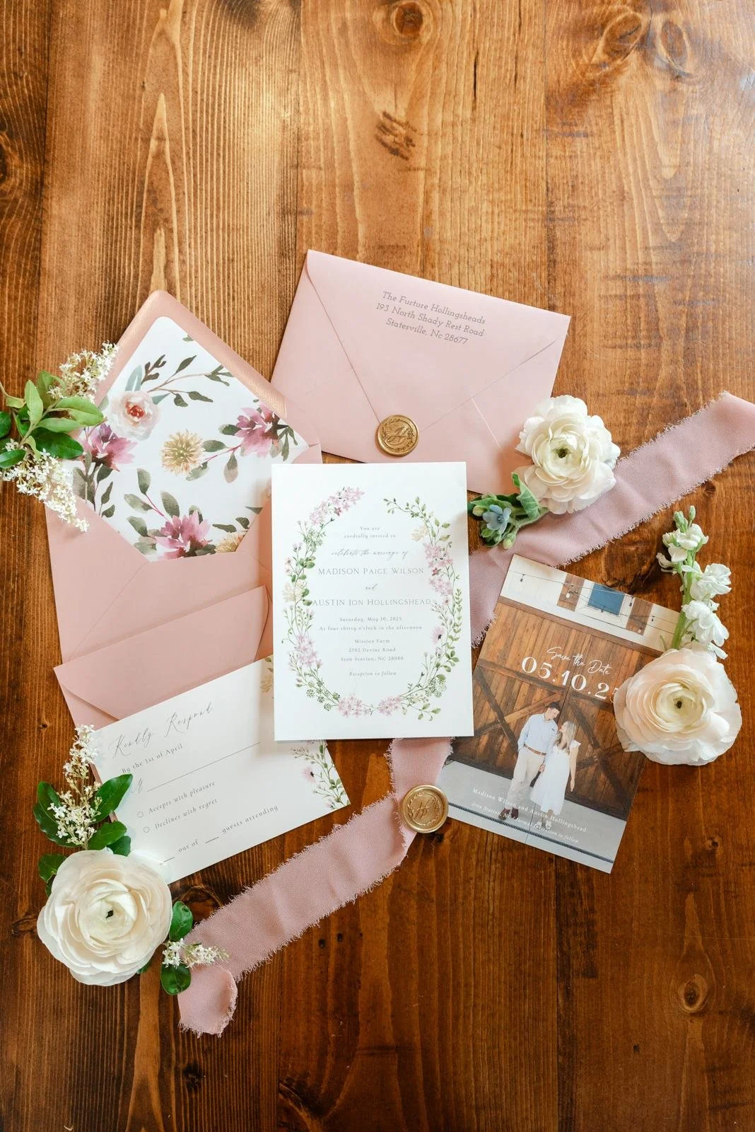 Wedding invitations and RSVP card with floral designs, pink envelopes, white flowers, a pink ribbon, and gold wax seal on a wooden surface.