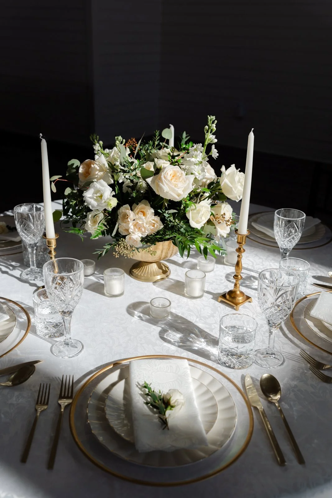 Elegant dining table with a floral centerpiece, gold-rimmed plates, gold cutlery, crystal glassware, white napkin with a small flower, and tall white candles in gold holders.