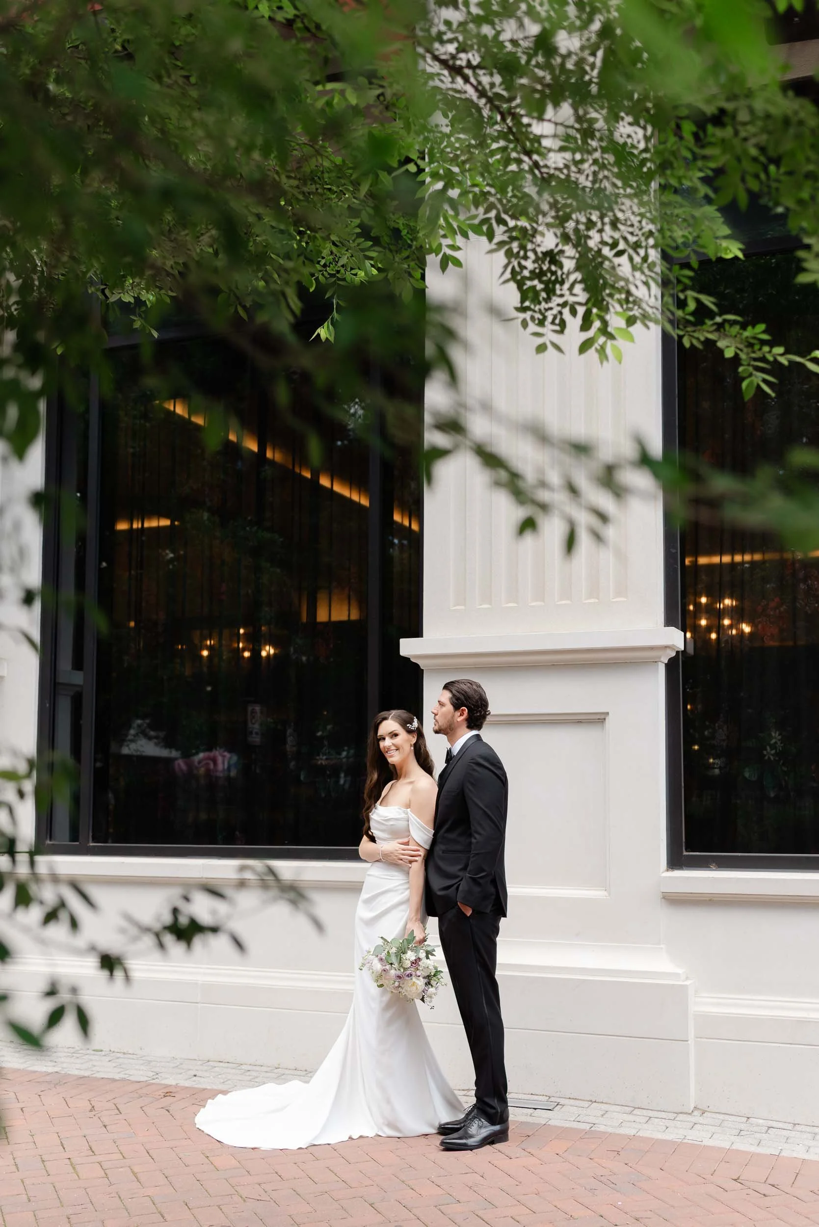 A newlywed couple standing outside in front of a modern building with large windows, partially obscured by green foliage. The bride is in a white wedding gown holding a bouquet, and the groom is in a black tuxedo. They are smiling and looking at each