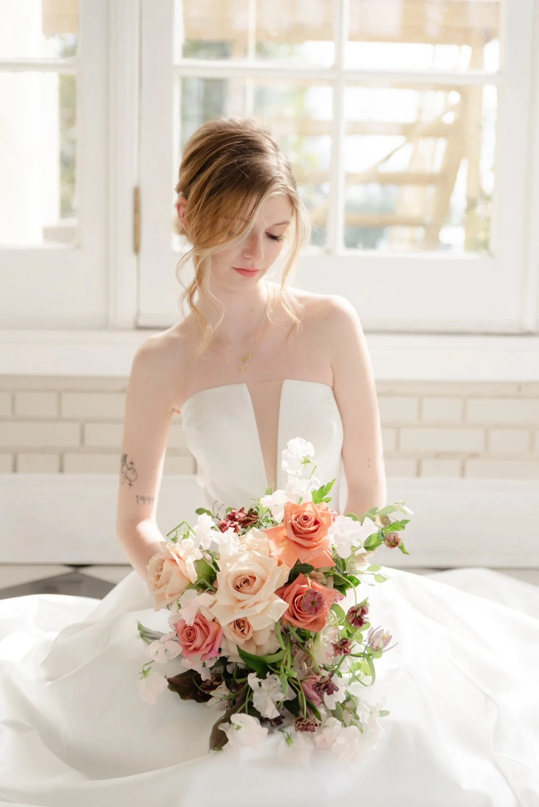 A woman in a white wedding dress with a deep V neckline sitting in front of a window holding a bouquet of pink and white roses and flowers.
