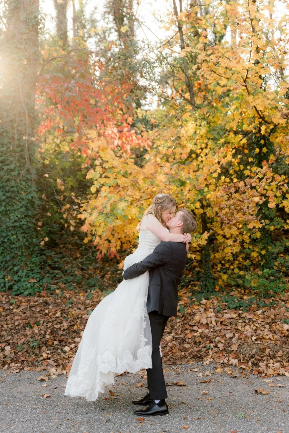 A man lifting a woman in a wedding dress outdoors surrounded by autumn leaves and colorful trees, during sunset.
