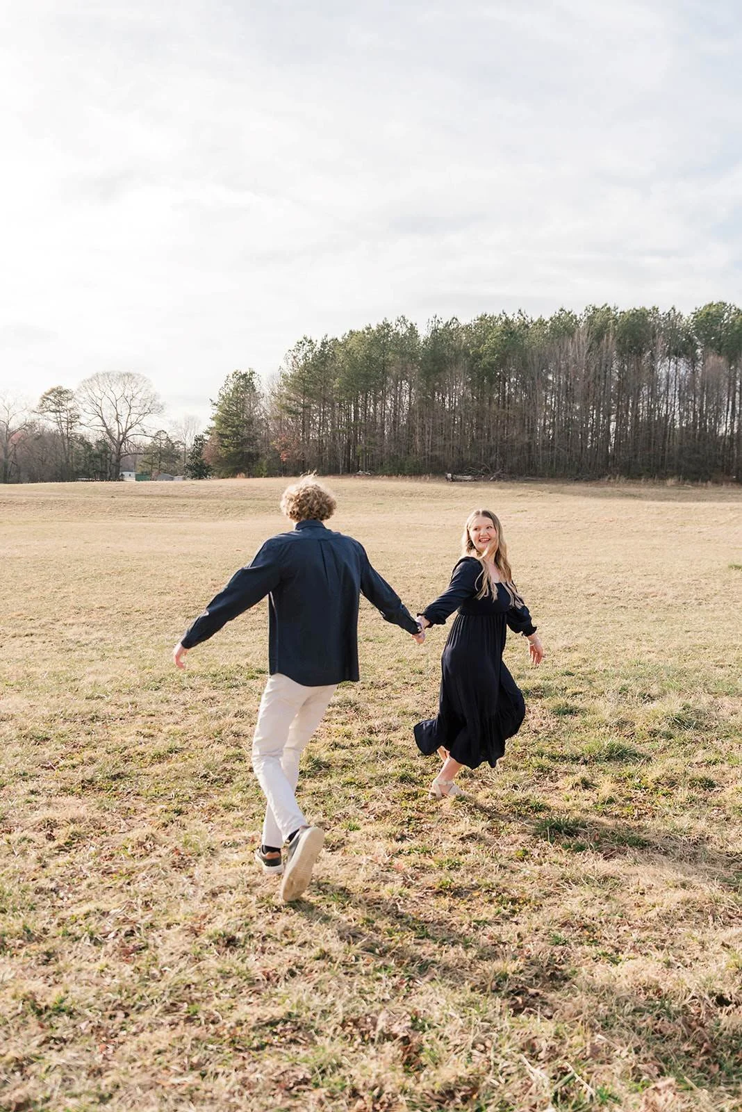 A couple holding hands and running through an open grassy field on a cloudy day, with trees in the background, the woman smiling and the man facing away from the camera.