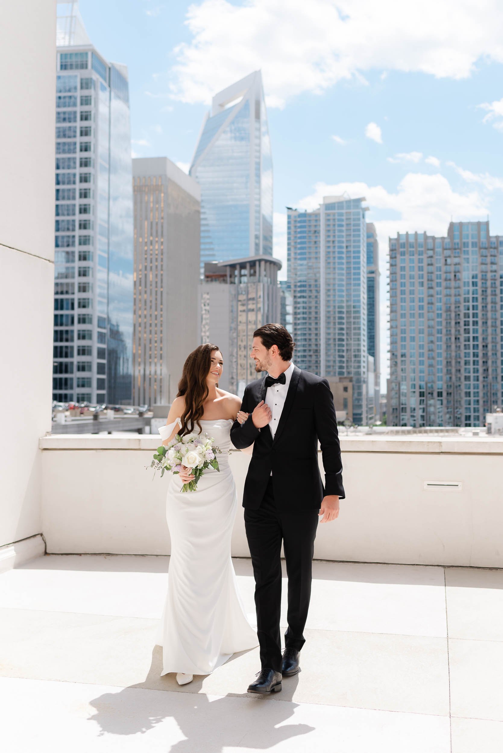 A bride in a white wedding gown holding a bouquet of flowers, walking arm-in-arm with a groom in a black tuxedo, on a rooftop with the Charlotte skyline background, including tall glass buildings under a partly cloudy sky.