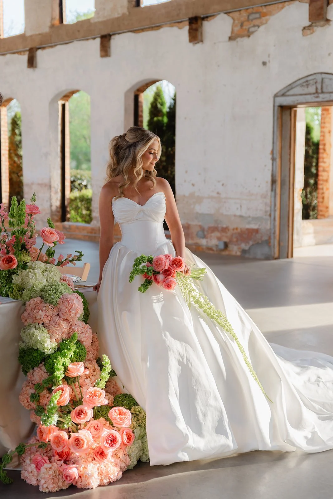 A bride in a white wedding gown sitting at a floral-decorated table inside a partially constructed building with exposed brick walls and open windows.