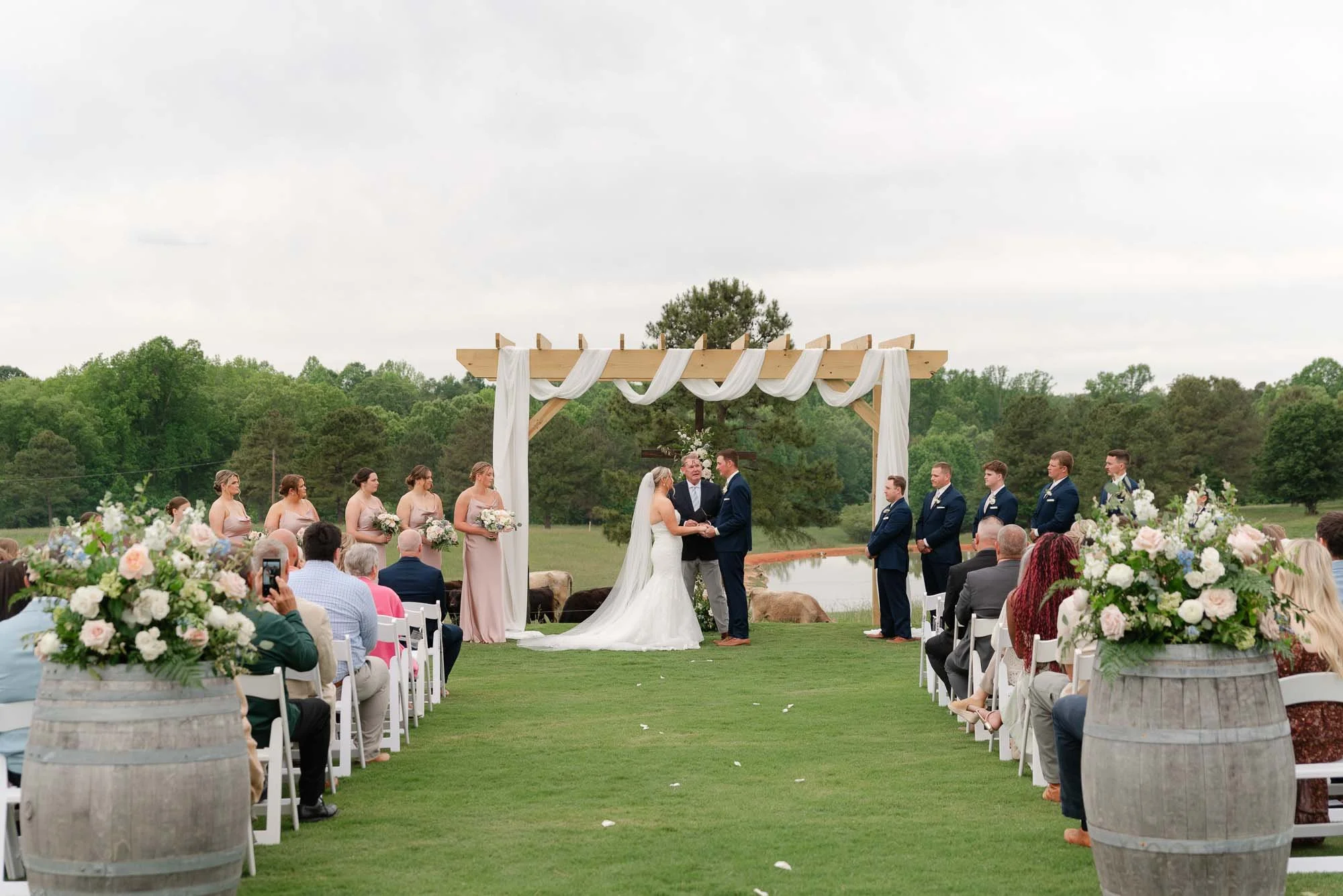 An outdoor wedding ceremony with a bride and groom exchanging vows under a wooden arch decorated with white fabric, surrounded by bridesmaids and groomsmen, with guests seated in front and large flower arrangements on barrels.