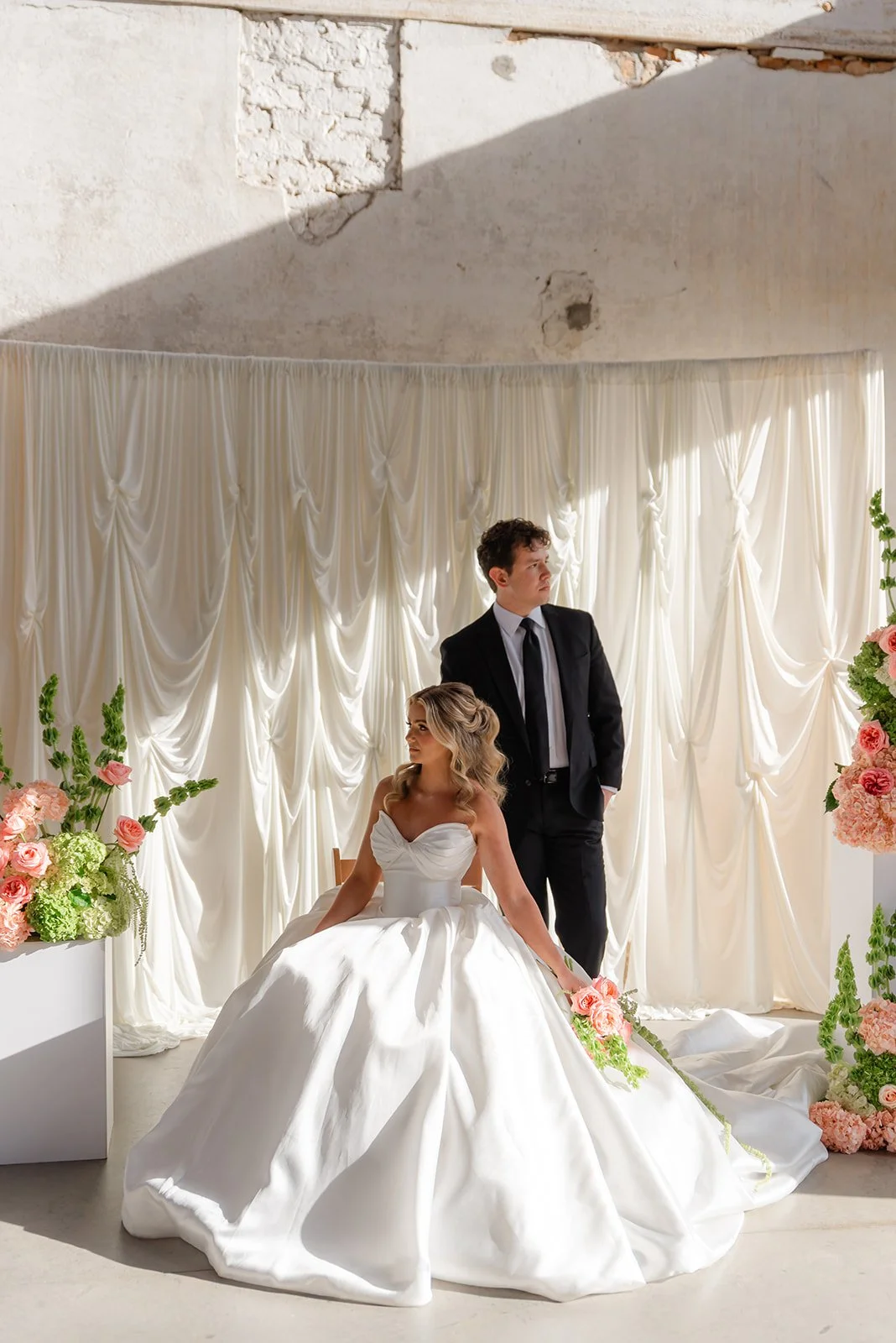 A bride in a white wedding gown sitting on a chair, next to a groom in a black suit, standing behind her, both in front of white drapery with floral arrangements around them.