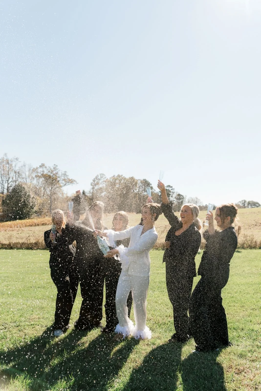 Group of women in formal attire celebrating outdoors with champagne, spraying it and laughing in a grassy field on a sunny day.