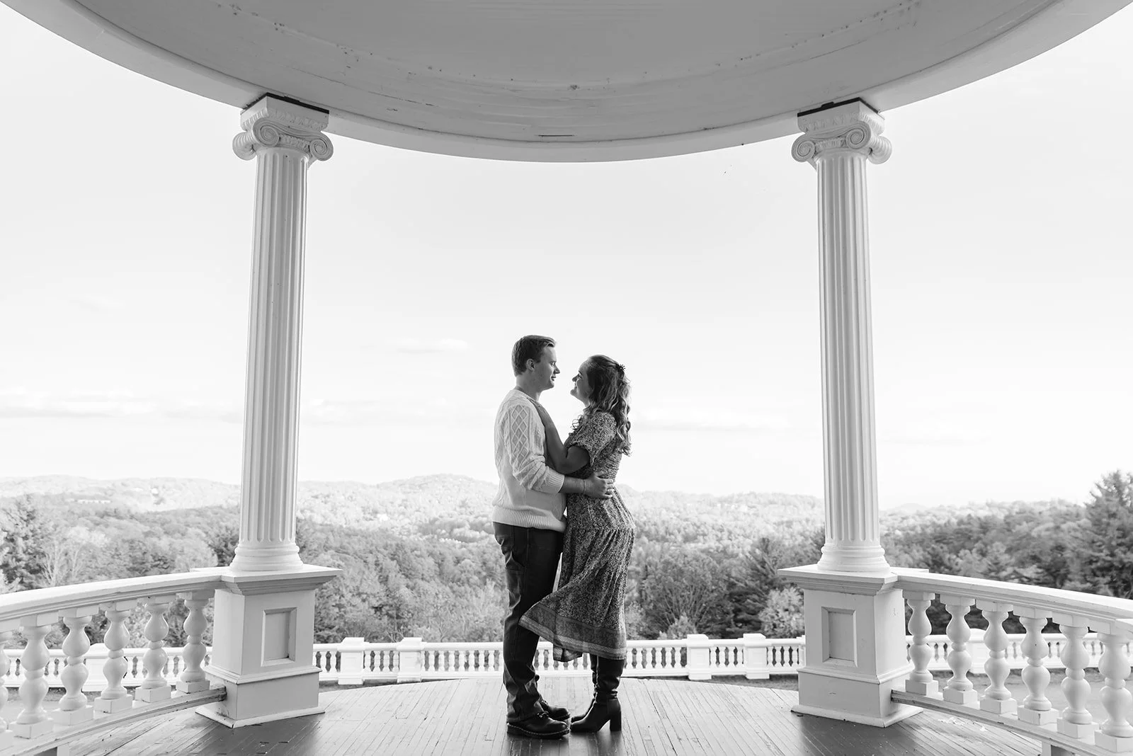 A couple standing close and looking at each other on a balcony with decorative columns, overlooking a scenic landscape of trees and hills in the background.