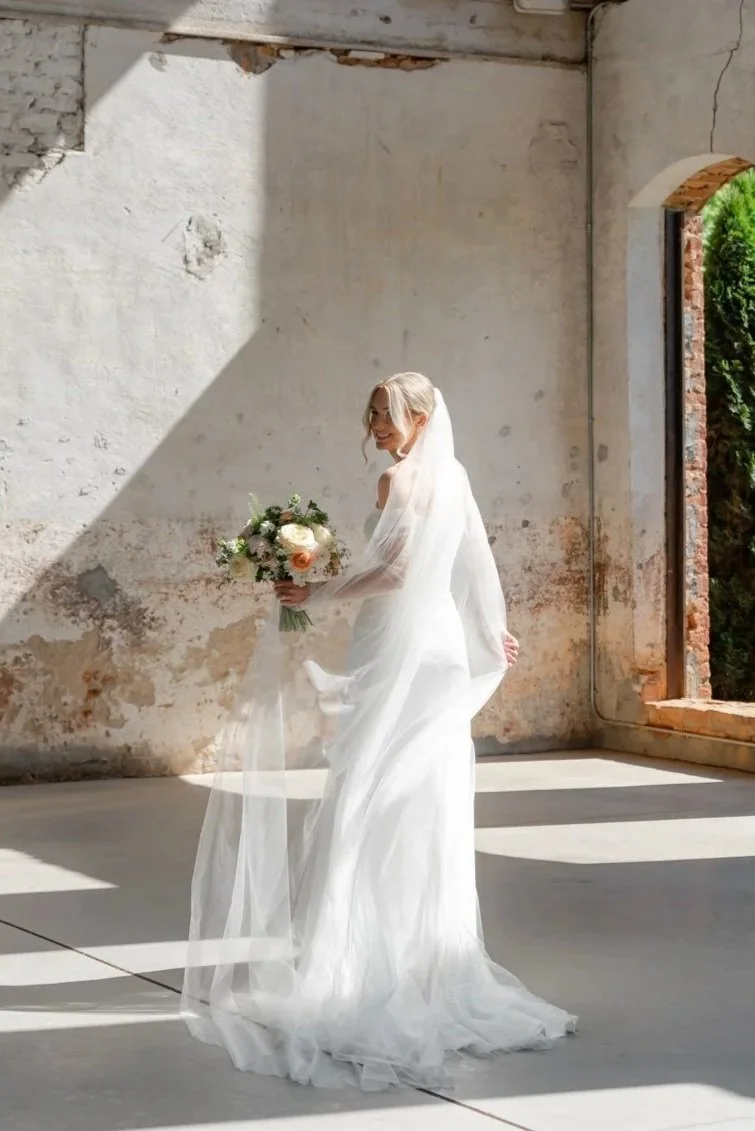 A bride wearing a white wedding gown and veil, holding a bouquet of flowers, standing in an industrial-style space with exposed brick and paint.