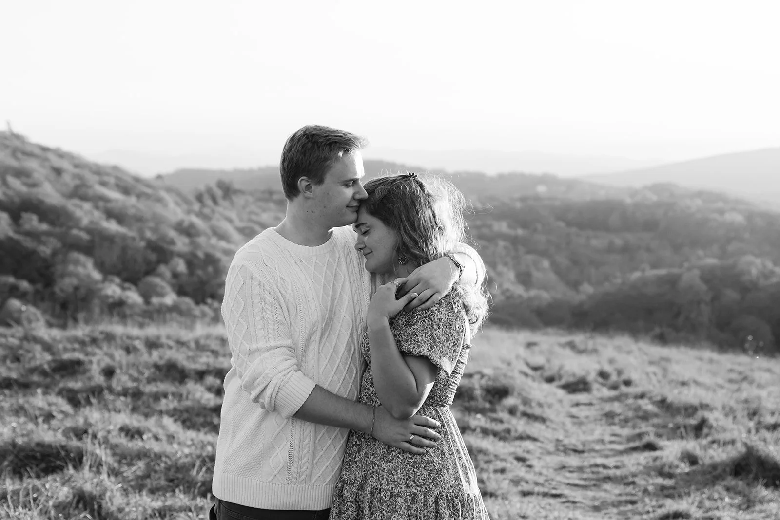 A black and white photo of a young couple embracing outdoors on a grassy hill, with rolling hills and trees in the background.