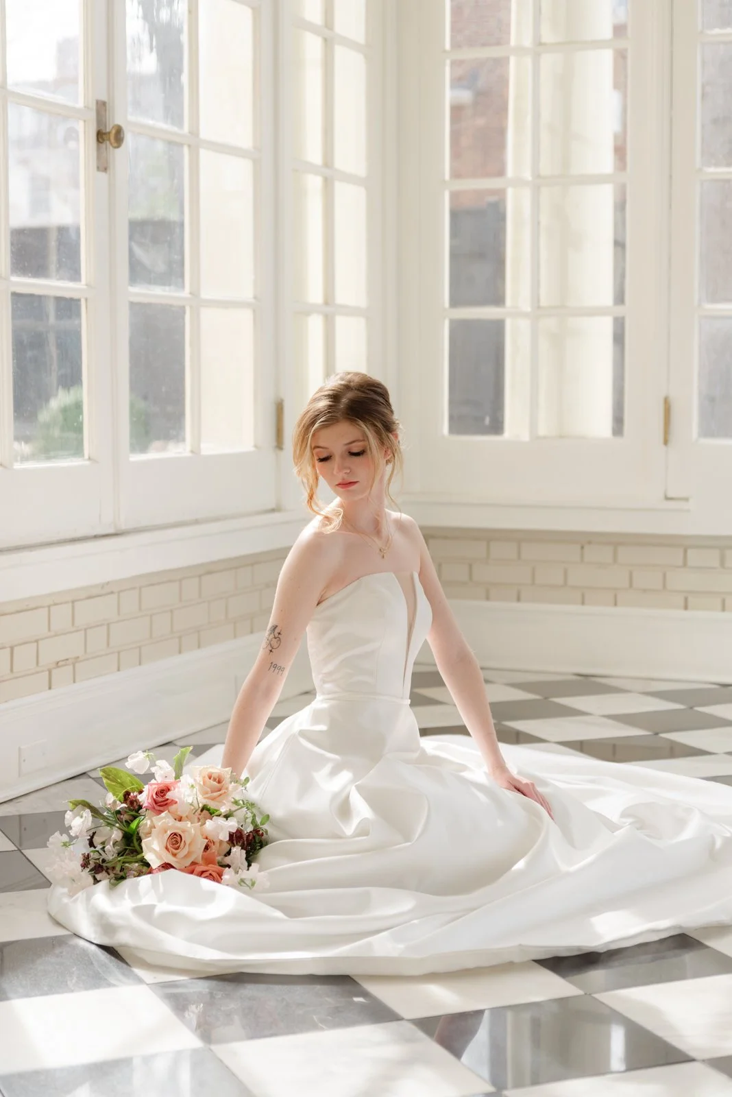 A woman in a white wedding dress sitting on a black and white checkered floor, holding a bouquet of pink and white roses, near large windows in a bright room with white brick walls.