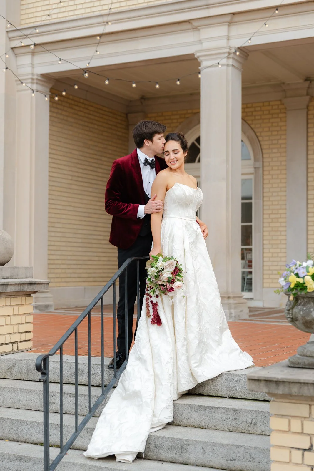 A bride in a white wedding gown holding a bouquet of flowers standing with a groom in a red velvet blazer and bow tie on the steps of Separk Mansion.