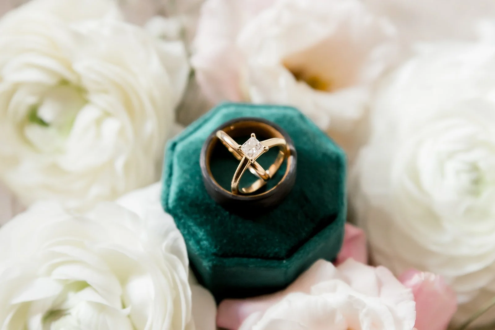 A gold ring with a square diamond sits in a green velvet ring box, surrounded by white and light pink flowers.
