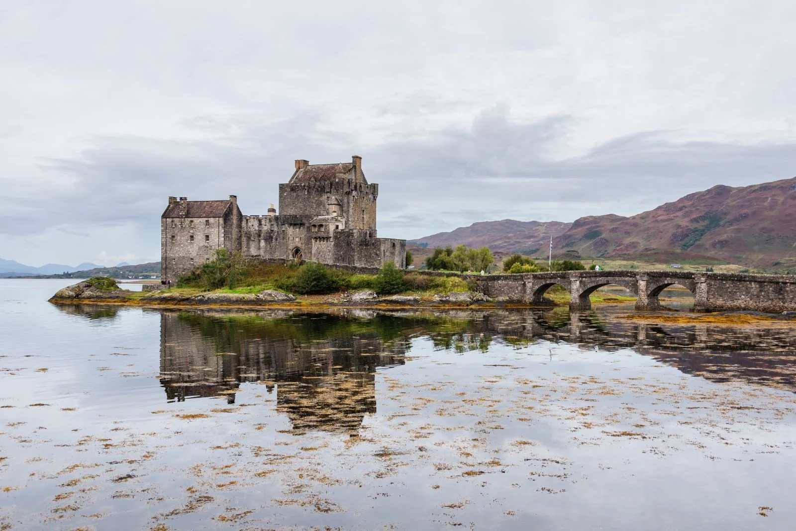 A medieval castle on a small island connected by a stone bridge, with calm water and mountain hills in the background, under an overcast sky.