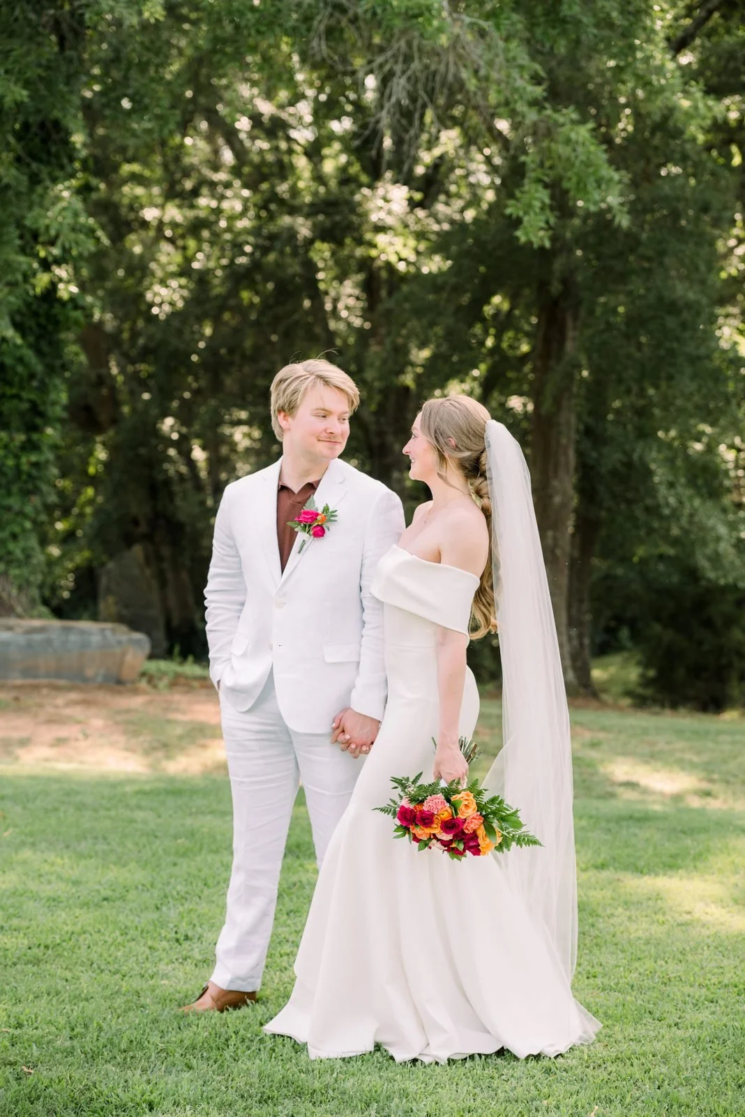 A bride and groom standing outdoors on a grassy area, holding hands and looking at each other, with trees in the background. The bride is in a white wedding gown with an off-shoulder design and a long veil, holding a colorful bouquet. The groom is in a white suit with a dark shirt and a boutonnière.