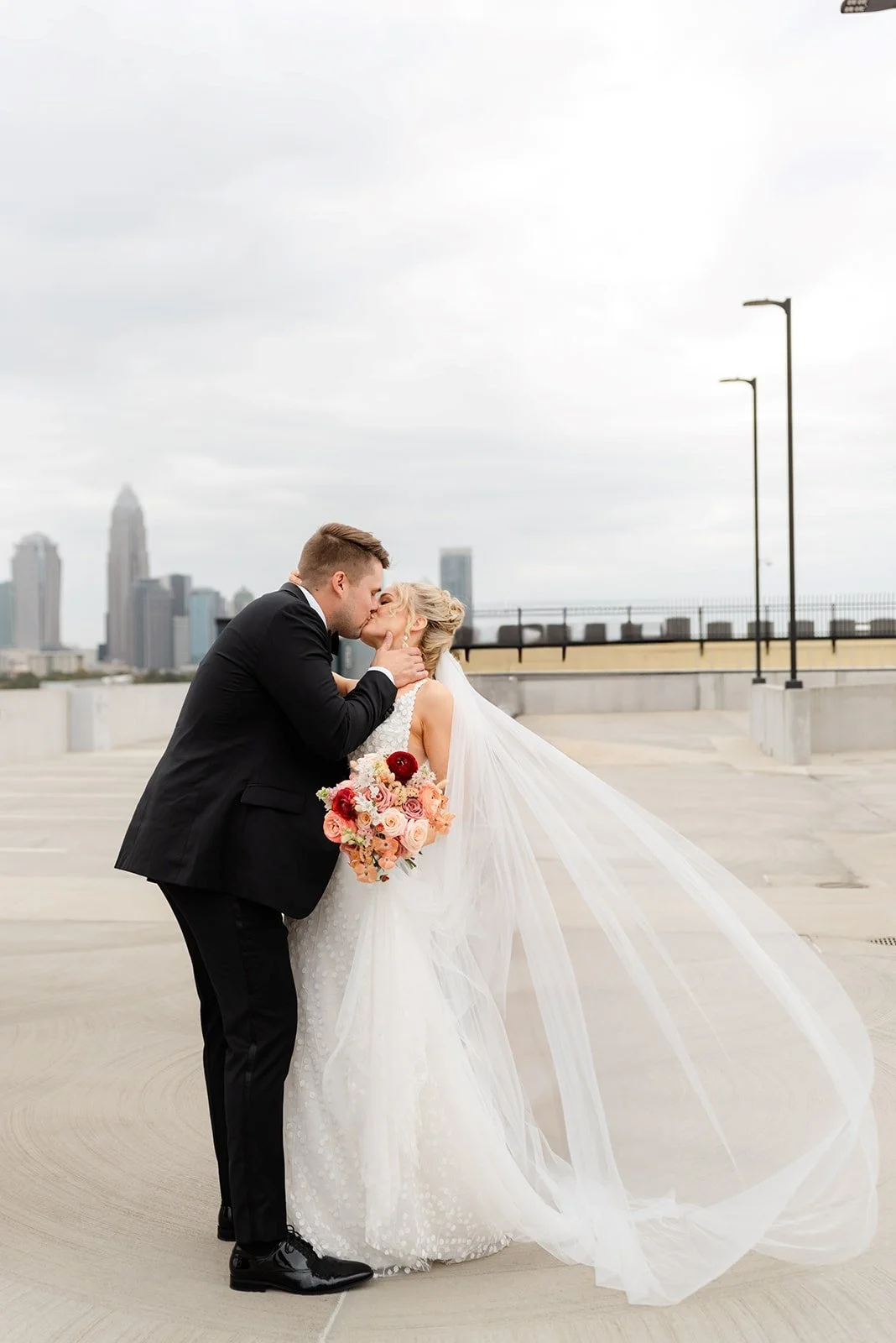 A bride and groom kissing on a rooftop with city skyline in the background, the bride holding a bouquet of pink and red flowers, and the groom dressed in a black suit.
