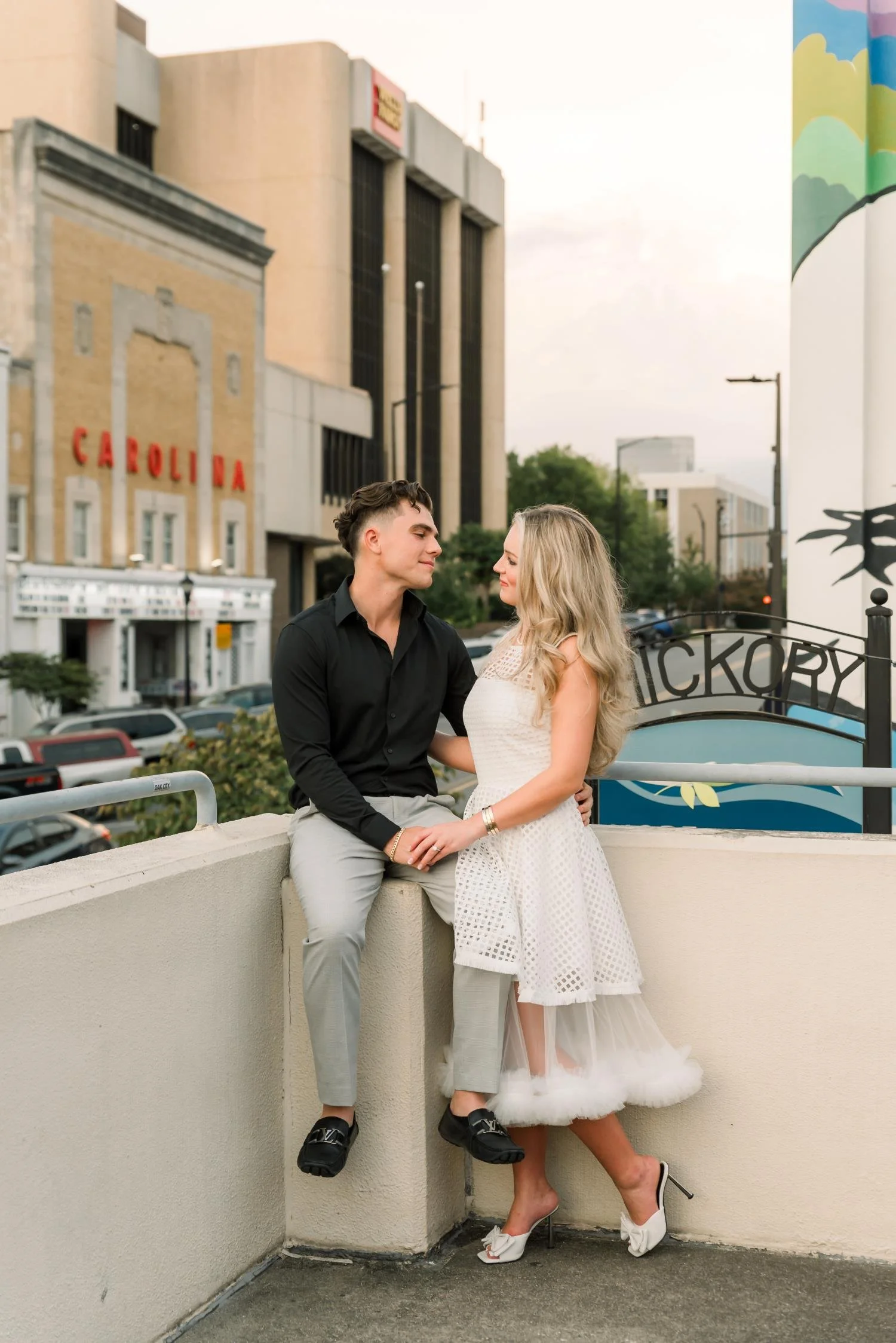 A young couple stands on a rooftop or balcony, with the young man sitting on the ledge and the woman standing close to him. They are facing each other affectionately, with the city street with parked cars, buildings, and a sign that reads 'CAROLINA' 