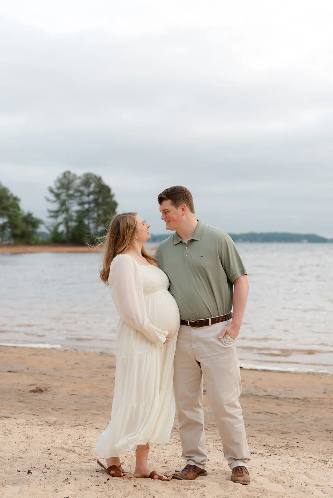 A pregnant woman in a cream-colored dress and a man in a green polo shirt and khakis standing on the beach, smiling and looking into each other's eyes.