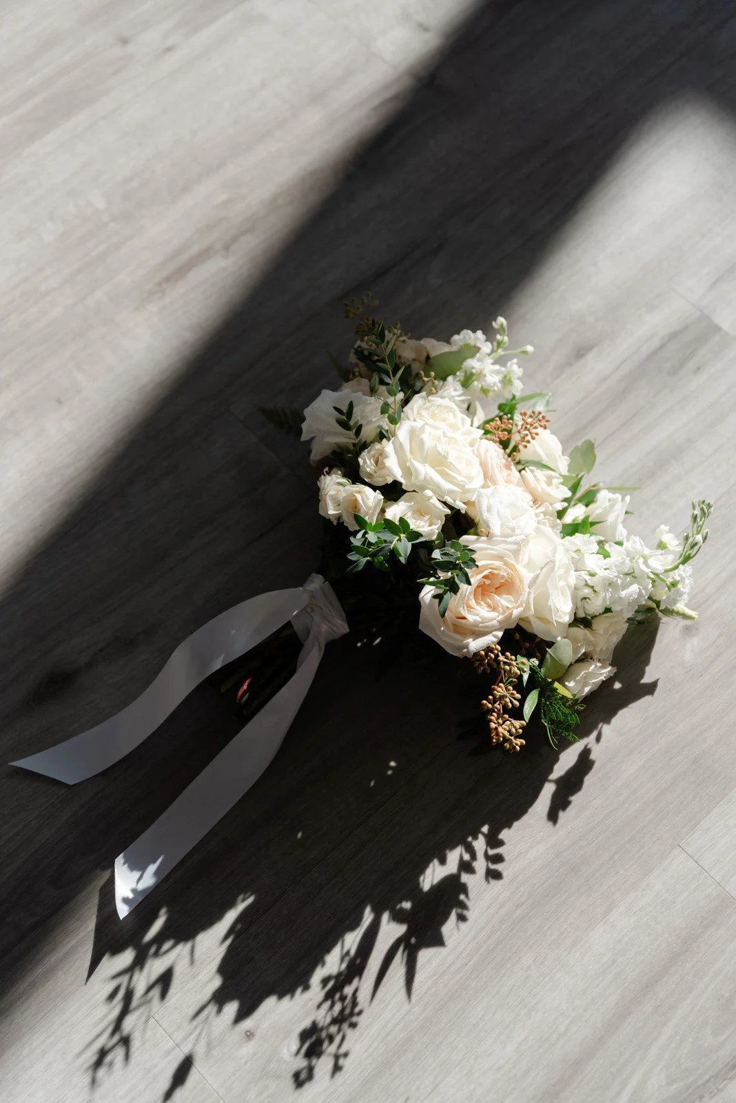 A bouquet of white flowers with greenery and a white ribbon, casting a shadow on a gray wooden floor.