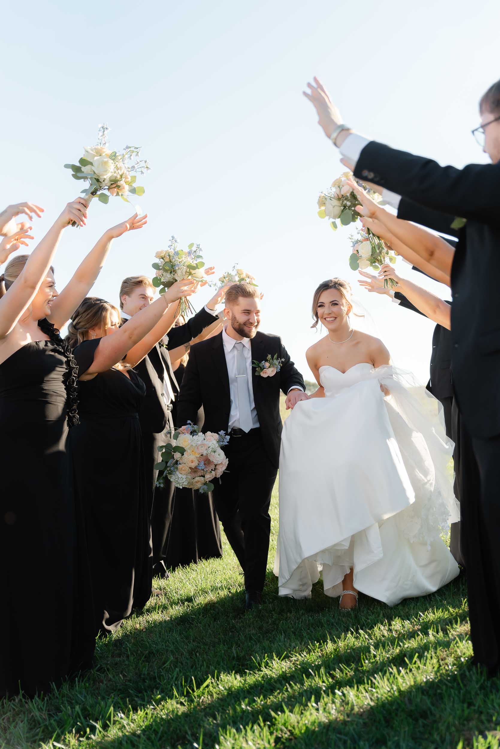 A wedding celebration outdoors with a bride and groom walking on grass, surrounded by guests holding bouquets and raising their hands, under a clear sky.
