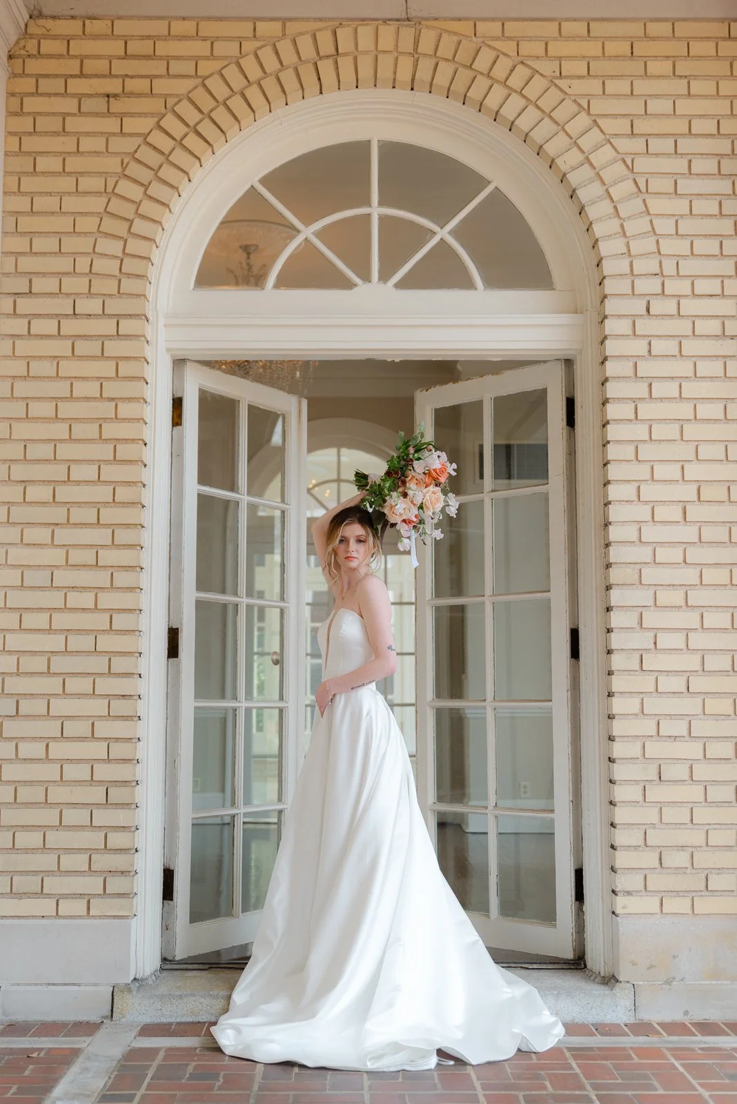 A woman in a wedding dress holding a bouquet of flowers above her head, standing in front of an open, double-set glass door with an arched window above, in a building with beige brick walls.