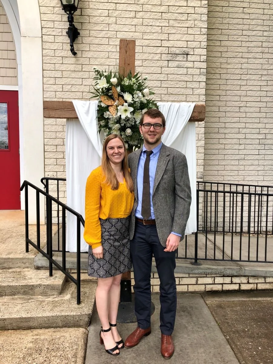 A smiling young woman and young man standing in front of a decorative floral arrangement with a cross backdrop during a church event. The woman is wearing a yellow blouse and patterned skirt, and the man is wearing a gray blazer, blue shirt, and dark pants.