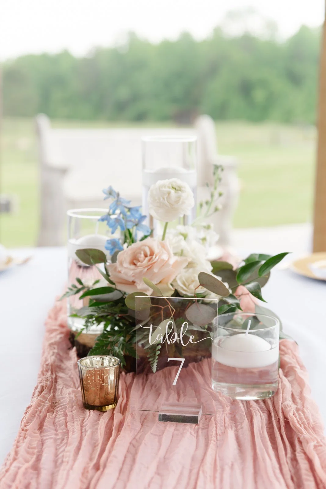 Elegant table centerpiece with pink and white flowers, green leaves, and candles on a pink table runner, with outdoor scenery in the background.